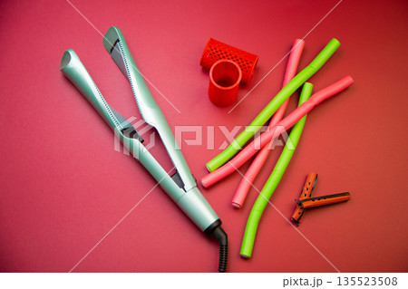 Top view of a green hair straightener with a twisted ceramic canvas, next to which there are curlers of different shapes and sizes on a pink background. The concept of styling, hairstyle, curls. High Top view of a green hair straightener with a twisted ceramic canvas, next to which there are curlers of different shapes and sizes on a pink background. The concept of styling, hairstyle, curls. High 135523508