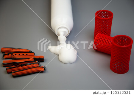 Side view of a bottle of foam for long-term hair styling, white foam poured out of it, next to it are pink curlers of different shapes and sizes on a gray background. The concept of styling, hairstyle 135523521