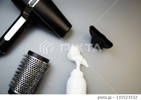 Close-up of hair styling foam in a white bottle, next to it a hairdryer with a round comb to create voluminous hairstyles on a gray background with space for text. The concept of long-term hair Close-up of hair styling foam in a white bottle, next to it a hairdryer with a round comb to create voluminous hairstyles on a gray background with space for text. The concept of long-term hair 135523532