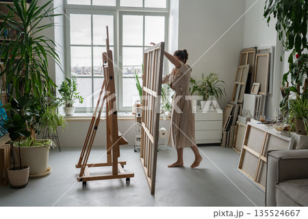 Focused woman painter artist finishing painting and taking canvas from easel at home workshop. 135524667