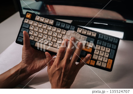 Man cleaning mechanical keyboard with disposable wipe, maintaining hygiene, ensuring germ-free surfaces, performing regular maintenance on office equipment to keep functional and clean. Man cleaning mechanical keyboard with disposable wipe, maintaining hygiene, ensuring germ-free surfaces, performing regular maintenance on office equipment to keep functional and clean. 135524707