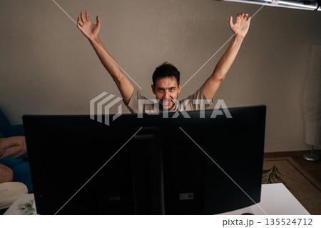 Young man reacting with excitement and joy, raising arms in celebration watching live broadcast on television, celebrating victory, cheering for favorable outcome, success at home, looking at camera. Young man reacting with excitement and joy, raising arms in celebration watching live broadcast on television, celebrating victory, cheering for favorable outcome, success at home, looking at camera. 135524712
