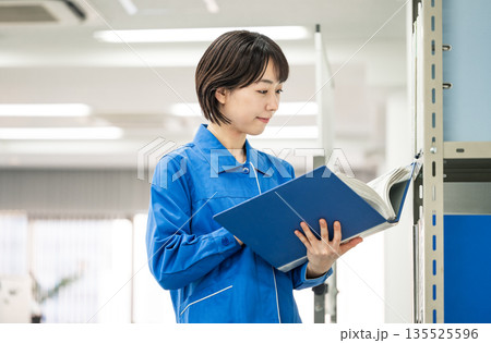 オフィスで資料を見る作業服姿の若い女性 オフィスで資料を見る作業服姿の若い女性 135525596