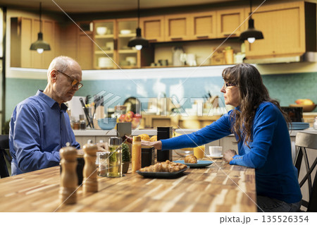 Mature woman asking her man to take a break from navigating on laptop during breakfast, closing the lid on device. Wife needing some attention while they drink coffee and serve food in the kitchen. Mature woman asking her man to take a break from navigating on laptop during breakfast, closing the lid on device. Wife needing some attention while they drink coffee and serve food in the kitchen. 135526354