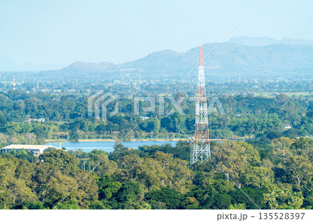 Large telecommunication tower standing tall in a lush green forest landscape with mountains and a lake in the background. Remote area signal coverage and rural connectivity infrastructure. 135528397