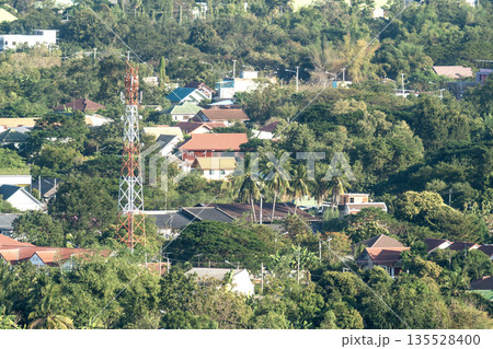 Tall orange and white telecommunications tower rising above a lush tropical town with colorful rooftops and green trees. High-angle view of communications infrastructure in a bright rural landscape. 135528400