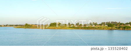 Wide panoramic view of a calm river with lush green riverbanks under a clear blue sky. A small boat cruises on the water near a rural shoreline with trees and distant buildings in the afternoon. Wide panoramic view of a calm river with lush green riverbanks under a clear blue sky. A small boat cruises on the water near a rural shoreline with trees and distant buildings in the afternoon. 135528402