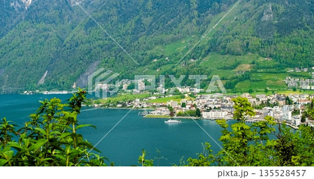 Enchanting Settlement Along Riverbank Surrounded By Vibrant Flora And Impressive Mountain Ranges 135528457