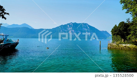 Sunlit Lake Surrounded By Mountains, Peaceful Alpine Scene With Boat And Vivid Blue Water Reflection 135528500