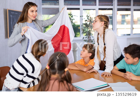 Smiling young female teacher explaining culture Japan while showing national flag to schoolchildren preteens in classroom 135529619
