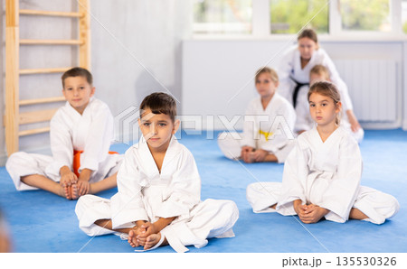 Little children in white kimono sits in a butterfly pose and practices stretching in gym 135530326