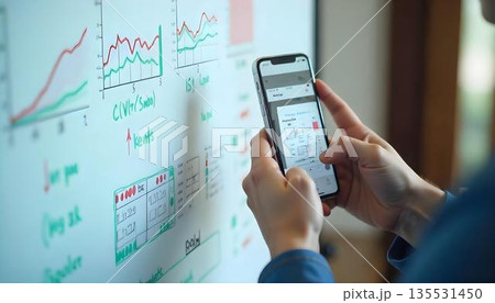 A suited individual studies a smartphone displaying financial graphs, emphasizing data-driven decision-making in a tech-savvy environment A suited individual studies a smartphone displaying financial graphs, emphasizing data-driven decision-making in a tech-savvy environment 135531450
