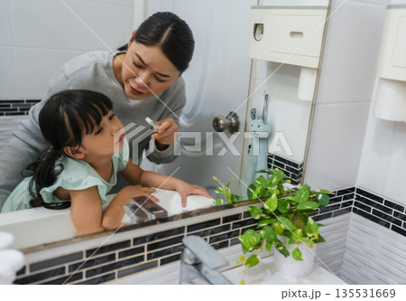 stubborn child girl refusing to brush her teeth in the bathroom. mother is trying to encourage her unhappy daughter during the morning routine, while child looks grumpy and resistant. 135531669