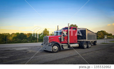 bold red semi truck with a long trailer sits in an open lot during sunset. The powerful rig highlights industrial design, chrome accents, and the rugged appeal of transportation and logistics 135532002