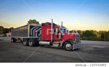 bold red semi truck with a long trailer sits in an open lot during sunset. The powerful rig highlights industrial design, chrome accents, and the rugged appeal of transportation and logistics 135532003
