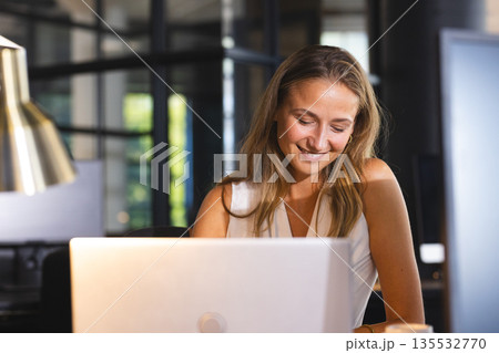 Smiling woman working on laptop at office during evening, feeling accomplished 135532770