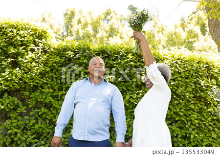 Senior couple enjoying sunny day outdoors, woman playfully holding bouquet up high Senior couple enjoying sunny day outdoors, woman playfully holding bouquet up high 135533049