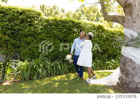 Senior couple embracing under tree in garden, enjoying romantic moment together 135533076