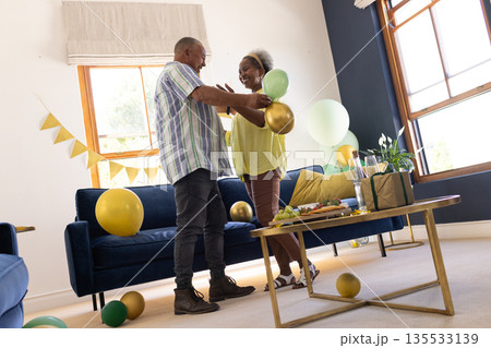 Senior couple dancing joyfully at home surrounded by colorful balloons Senior couple dancing joyfully at home surrounded by colorful balloons 135533139