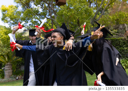 Graduating teens celebrating outdoors, wearing caps and gowns, holding diplomas Graduating teens celebrating outdoors, wearing caps and gowns, holding diplomas 135533835