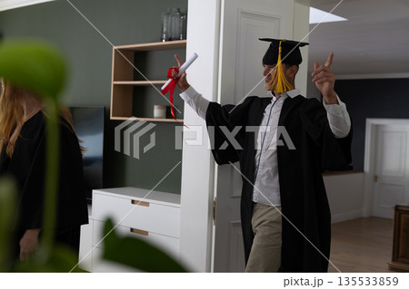Teen celebrating graduation at home, wearing cap and gown, holding diploma 135533859