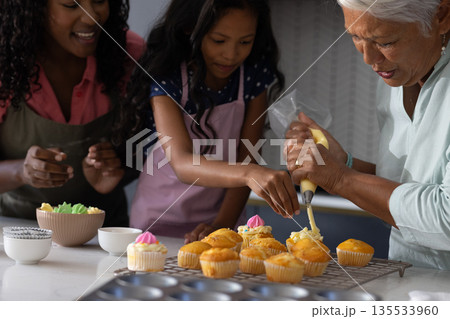 In kitchen, three generations decorating cupcakes, sharing family bonding time together 135533960