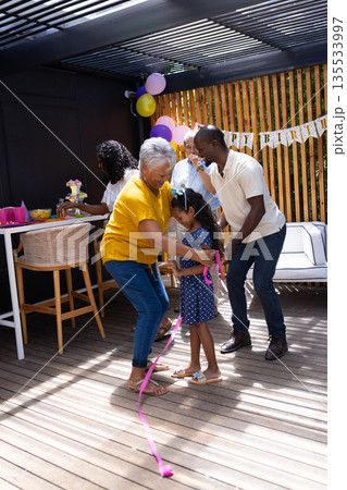 Grandmother and father helping girl with ribbon at outdoor birthday celebration Grandmother and father helping girl with ribbon at outdoor birthday celebration 135533997
