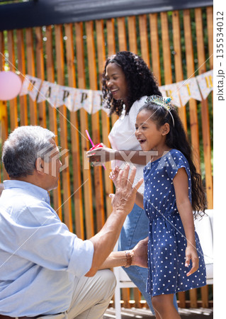 Grandfather playing with granddaughter, mother smiling at outdoor birthday party 135534012