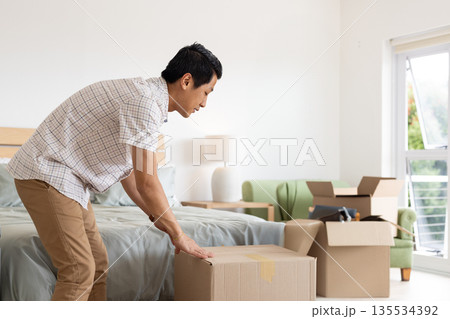 Mid adult Asian man bending over lifting cardboard box in bedroom with wooden headboard and lamp Mid adult Asian man bending over lifting cardboard box in bedroom with wooden headboard and lamp 135534392