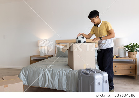 Asian man unpacking soccer ball from cardboard box in bedroom with rolling suitcase, copy space 135534396