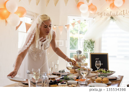 Bride wearing lace gown and veil arranging dishes on table in indoor reception with peach balloons Bride wearing lace gown and veil arranging dishes on table in indoor reception with peach balloons 135534398