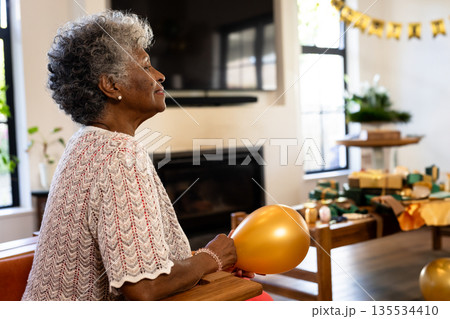 Senior woman sitting at wooden table in living room, holding gold balloon and gifts, copy space Senior woman sitting at wooden table in living room, holding gold balloon and gifts, copy space 135534410