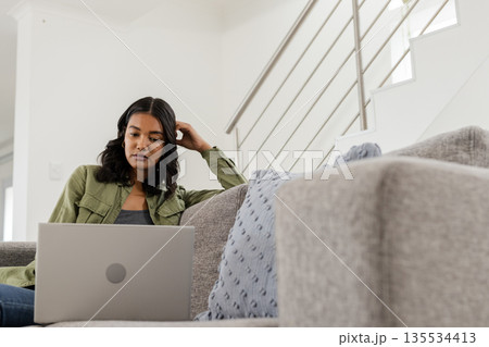 African American woman sitting on gray sofa leaning against cushion using laptop by metal staircase African American woman sitting on gray sofa leaning against cushion using laptop by metal staircase 135534413
