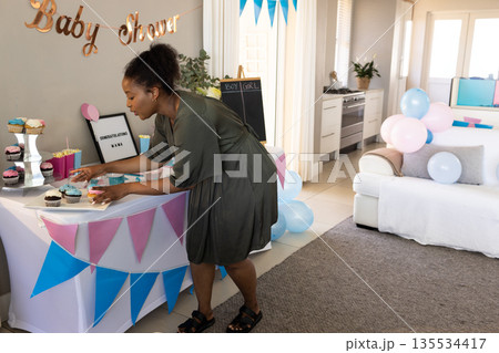 African American pregnant woman arranging cupcakes in living room, with baby shower banner African American pregnant woman arranging cupcakes in living room, with baby shower banner 135534417