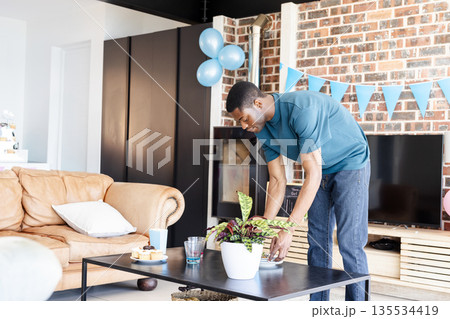 Mid adult African American man arranging potted plant on coffee table in living room, copy space Mid adult African American man arranging potted plant on coffee table in living room, copy space 135534419