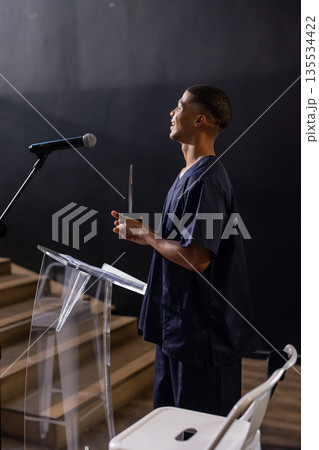 Man standing behind podium with microphone on stage holding trophy smiling upward, copy space 135534422