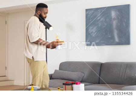 African American man celebrating birthday holding cake in modern living room with gifts, party hat African American man celebrating birthday holding cake in modern living room with gifts, party hat 135534426