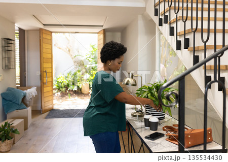African American man adjusting potted plant on console table in foyer with coffee cups, copy space 135534430