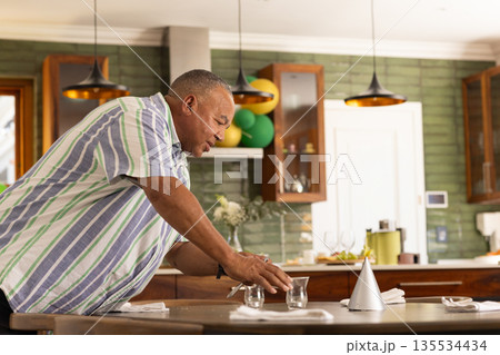 Senior man placing party hat and glasses on dining table in kitchen with balloons, copy space Senior man placing party hat and glasses on dining table in kitchen with balloons, copy space 135534434