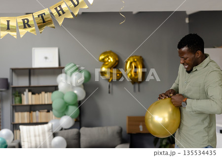 Mid-adult African American man holding gold balloon at home with foil 20 party balloons, copy space 135534435