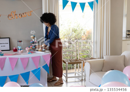 African American woman arranging cupcakes on cupcake stand in living room with bunting, copy space 135534456