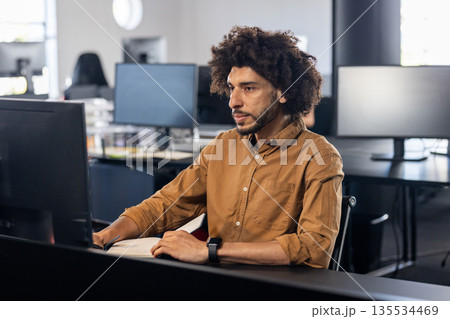 Young adult Asian man typing code at desk in open?plan office, with computer monitor and smartwatch 135534469