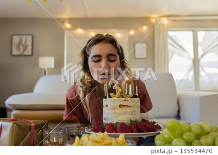 Female leaning forward and blowing out cake candles in living room, with gift, chips and grapes 135534470