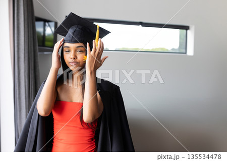 Woman adjusting mortarboard cap indoors in modern room, wearing graduation gown over red dress 135534478