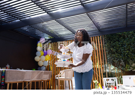 African American woman arranging cupcakes on stand under backyard pergola, with balloon garland African American woman arranging cupcakes on stand under backyard pergola, with balloon garland 135534479