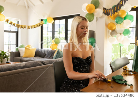 Woman sitting at table unwrapping birthday gift in living room with balloons, banner, copy space Woman sitting at table unwrapping birthday gift in living room with balloons, banner, copy space 135534490