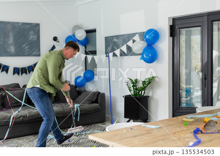 middle-aged man sweeping floor with broom in living room, clearing paper streamers and balloons 135534503