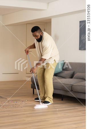 African American man sweeping colorful confetti off light wood floor in living room, with broom 135534506
