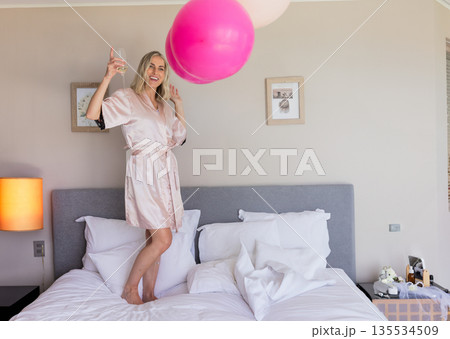Young woman standing on bed in modern bedroom, celebrating with pink balloons and wine glass 135534509