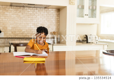 African American child girl writing in notebook in home kitchen at table with pencil, copy space African American child girl writing in notebook in home kitchen at table with pencil, copy space 135534514